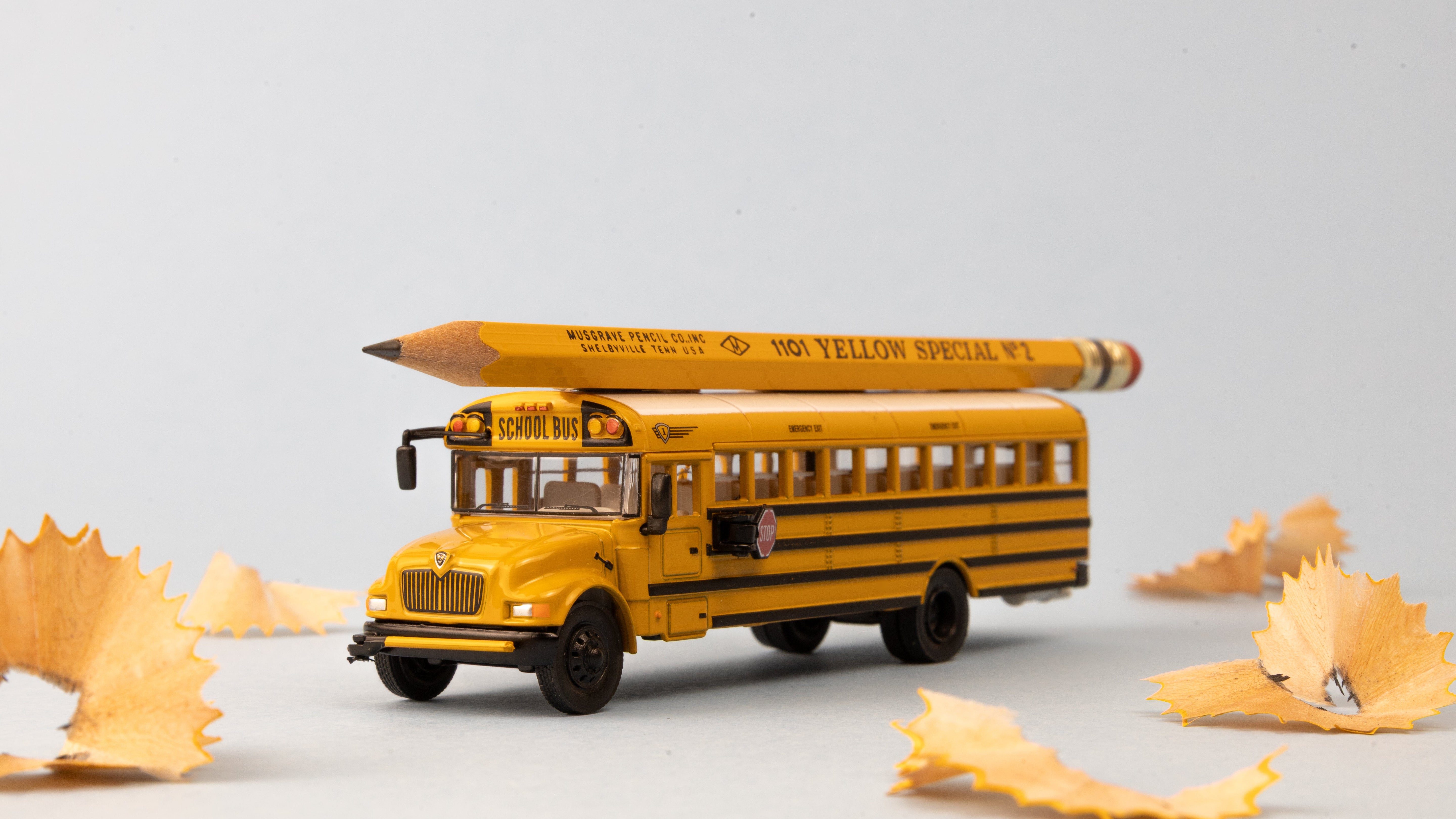 Model yellow school bus with pencil on top surrounded by pencil shavings on a light gray background